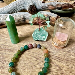 May include: A green jade stone bracelet with multi-colored agate beads. The bracelet is displayed on a wooden surface with a green jade stone, a green crystal tree, and a glass jar with a pink rose quartz crystal inside.