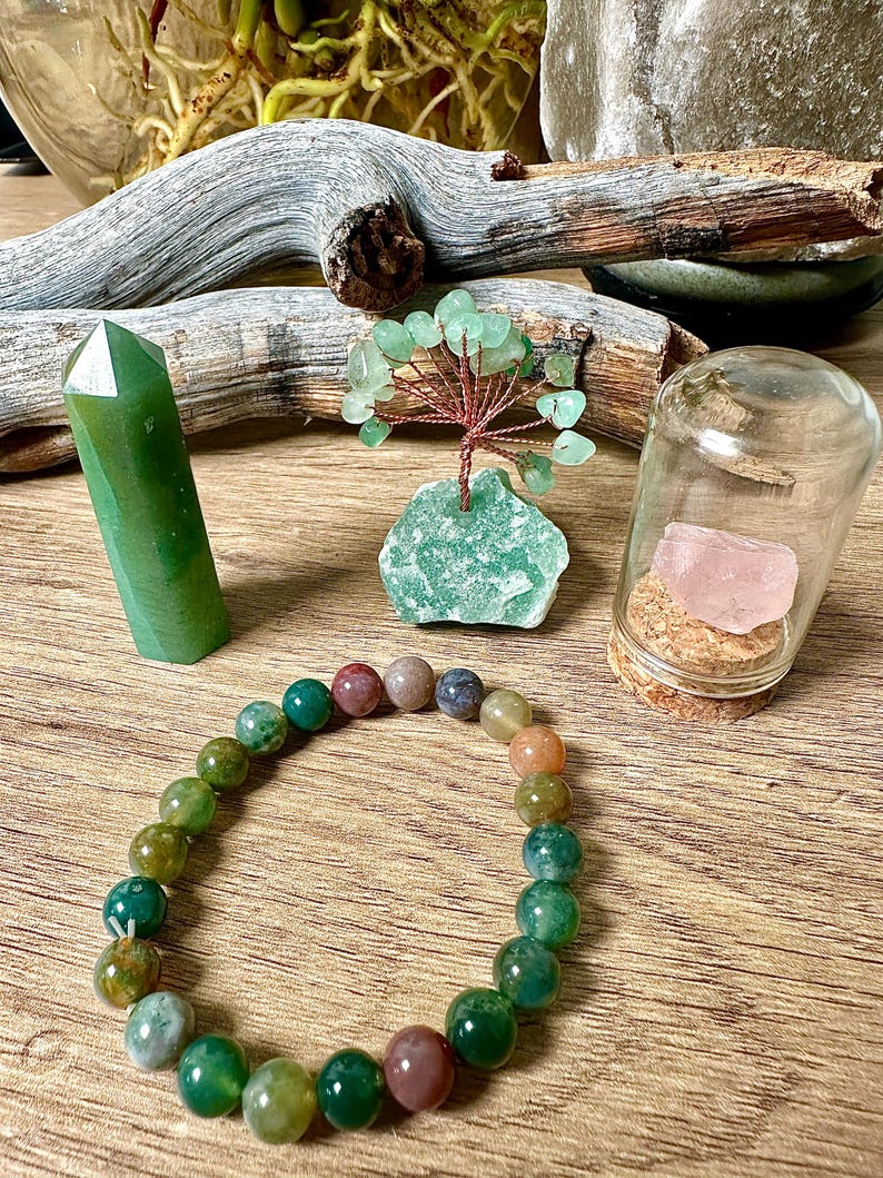 May include: A green jade stone bracelet with multi-colored agate beads. The bracelet is displayed on a wooden surface with a green jade stone, a green crystal tree, and a glass jar with a pink rose quartz crystal inside.
