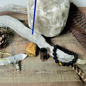 May include: A blue incense stick burning on a brown stone holder, surrounded by a black beaded bracelet, a white crystal, a brown and white crystal pendant, and a natural brown branch.