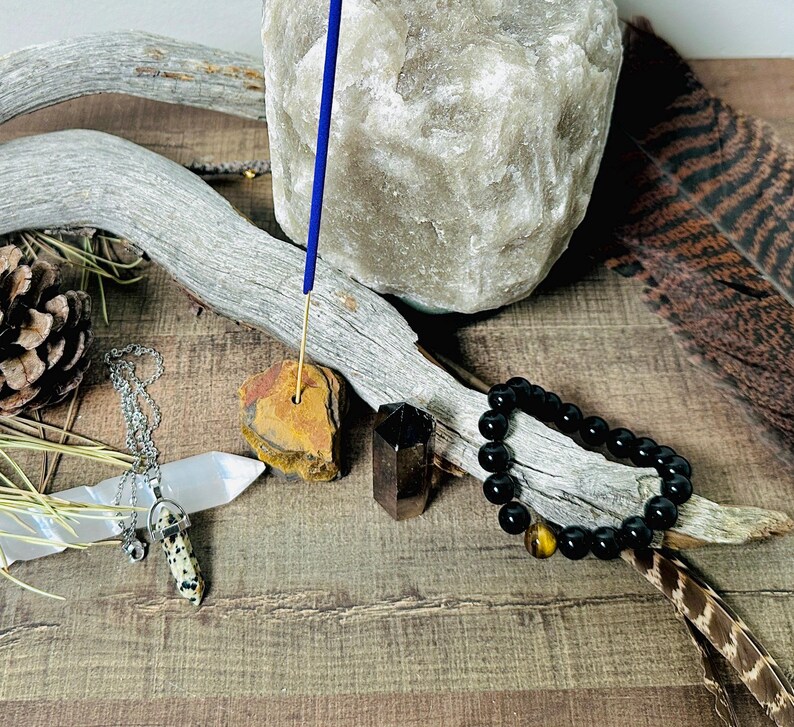 May include: A blue incense stick burning on a brown stone holder, surrounded by a black beaded bracelet, a white crystal, a brown and white crystal pendant, and a natural brown branch.