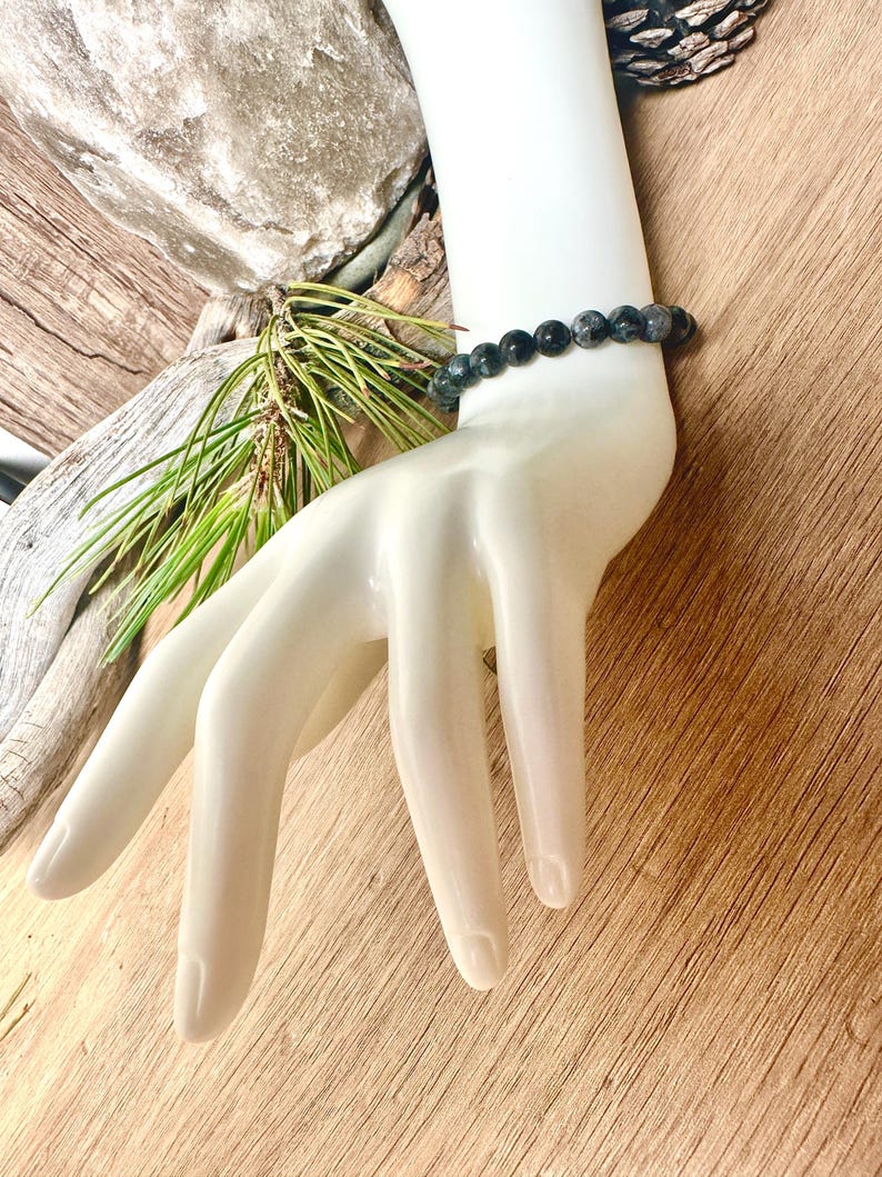 May include: A black and grey beaded bracelet on a white mannequin hand. The bracelet is made of round beads and is about 7 inches long.