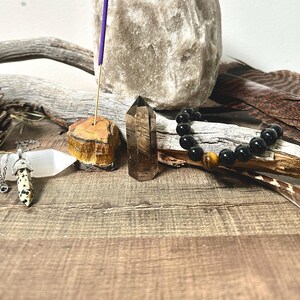 Puede incluir: Un conjunto de curación con cristales que incluye una punta de cuarzo transparente, un porta incienso de ojo de tigre, una varita de selenita blanca, un brazalete de ónix negro con una cuenta de ojo de tigre y un palo de incienso morado.