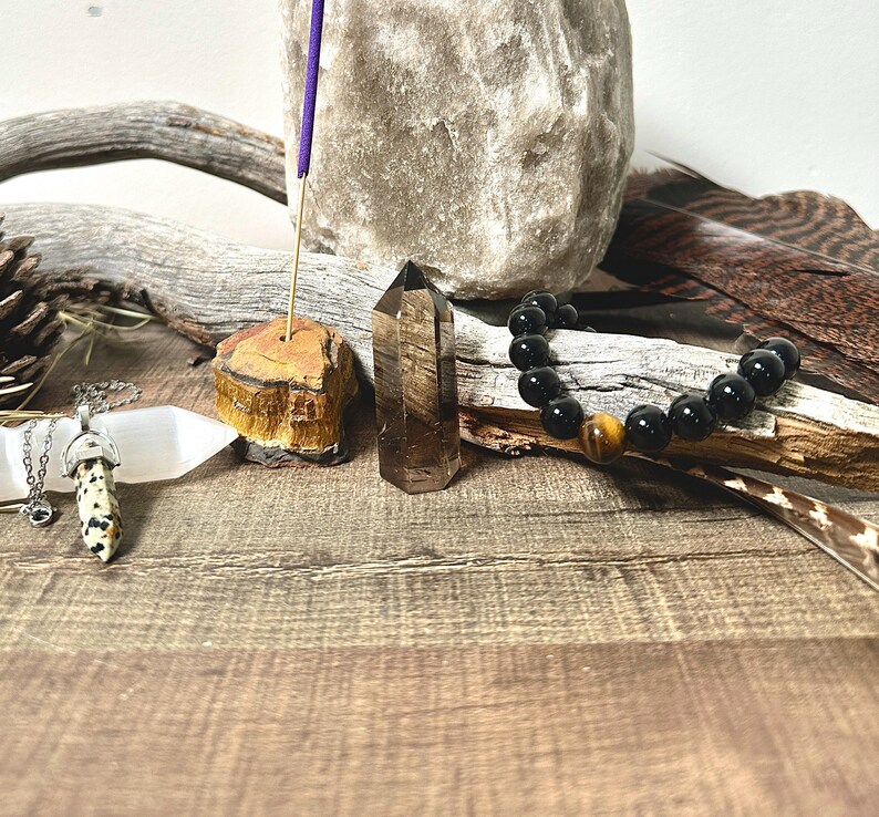 May include: A crystal healing set with a clear quartz point, a tiger's eye incense holder, a white selenite wand, a black onyx bracelet with a tiger's eye bead, and a purple incense stick.