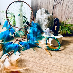 May include: A blue and green beaded bracelet, a black crystal with gold moon phases, a blue star-shaped crystal, and a dreamcatcher with blue and white feathers in a wooden box.