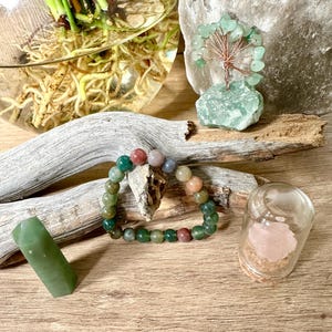 May include: A multi-colored agate beaded bracelet with green, red, and brown beads. The bracelet is on a wooden surface with a small glass jar containing a pink crystal, a green crystal, and a green wire tree.
