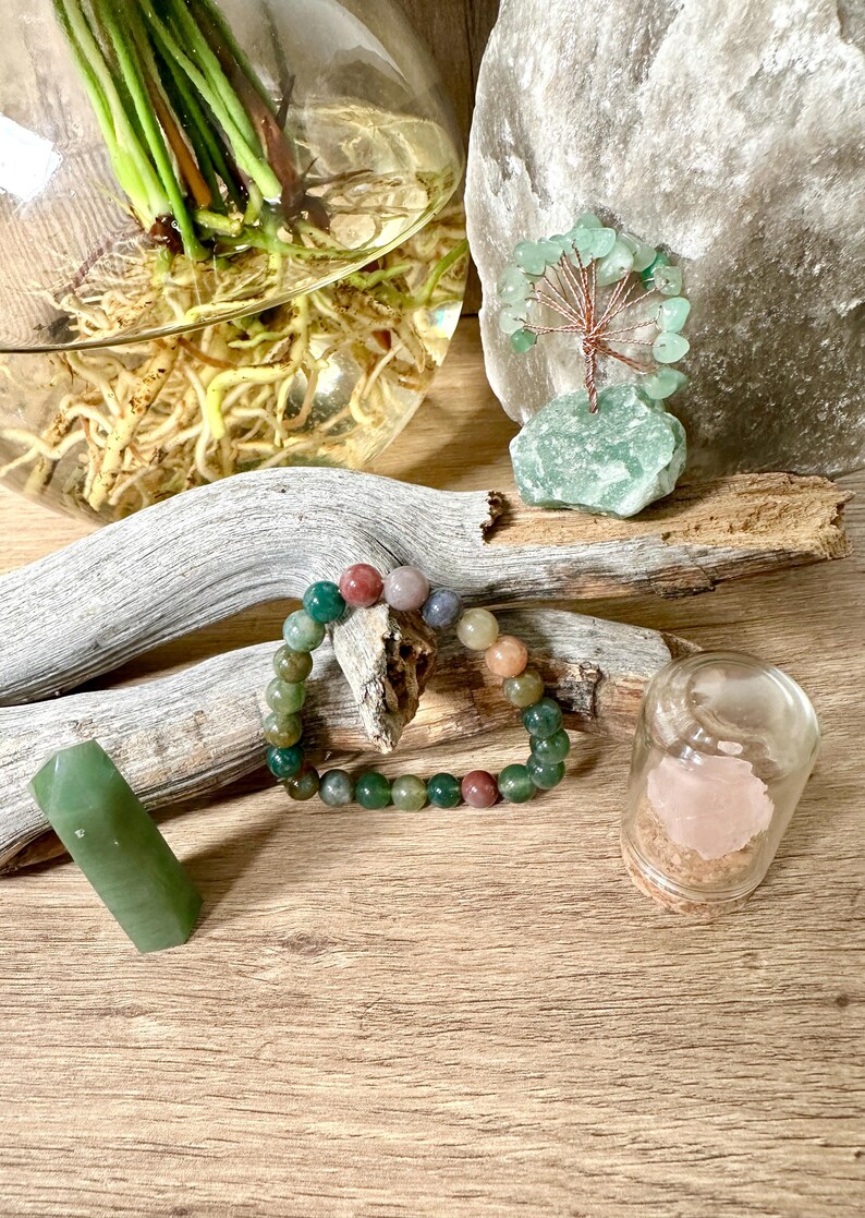 May include: A multi-colored agate beaded bracelet with green, red, and brown beads. The bracelet is on a wooden surface with a small glass jar containing a pink crystal, a green crystal, and a green wire tree.