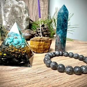 May include: A crystal pyramid with a turquoise tree inside, a blue crystal point, a brown tiger's eye stone, and a grey beaded bracelet.