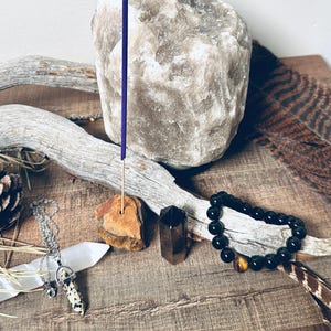 May include: A collection of spiritual and healing items including a Himalayan salt rock, a wooden incense holder with a purple incense stick, a black beaded bracelet with a tiger's eye bead, a white crystal, and a brown and white crystal pendant on a silver chain.