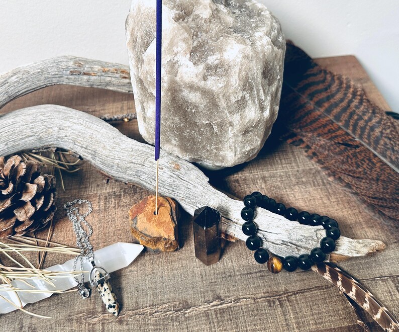 May include: A collection of spiritual and healing items including a Himalayan salt rock, a wooden incense holder with a purple incense stick, a black beaded bracelet with a tiger's eye bead, a white crystal, and a brown and white crystal pendant on a silver chain.