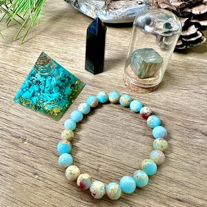 May include: A turquoise and gold crystal pyramid, a black crystal point, a clear glass jar with a cork lid containing a silver-colored crystal, and a light blue and white beaded bracelet on a wooden surface.