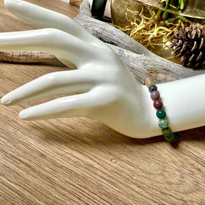May include: A bracelet made of round, multi-colored gemstone beads. The beads are shades of green, brown, pink, and purple. The bracelet is on a white mannequin wrist.