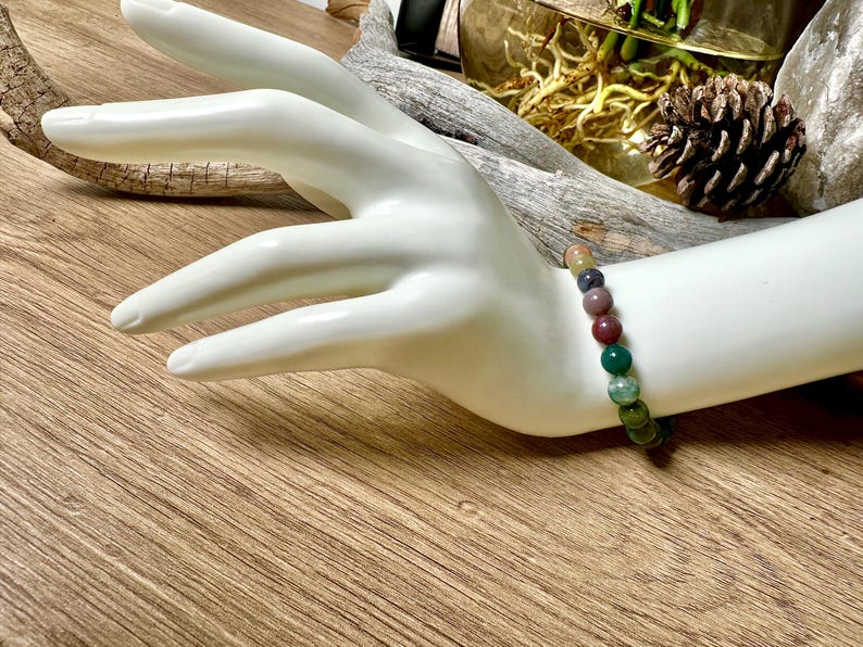 May include: A bracelet made of round, multi-colored gemstone beads. The beads are shades of green, brown, pink, and purple. The bracelet is on a white mannequin wrist.