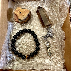 May include: A box filled with bubble wrap containing a black beaded bracelet with a tiger's eye bead, a brown crystal point, a small brown rock with a hole in it, and a silver chain with a black and white stone pendant.
