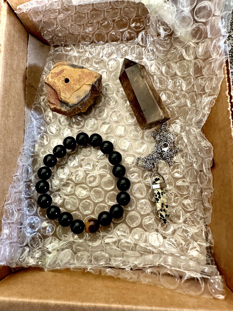 May include: A box filled with bubble wrap containing a black beaded bracelet with a tiger's eye bead, a brown crystal point, a small brown rock with a hole in it, and a silver chain with a black and white stone pendant.