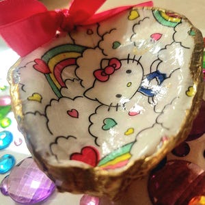 Puede incluir: Un cuenco decorativo de concha marina pintado a mano con un diseño de Hello Kitty con un arcoíris, nubes y corazones. La concha tiene un borde dorado y un lazo de cinta roja. El cuenco está rodeado de gemas de colores.