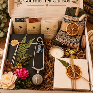 May include: A gift box labeled Book Lover & Gourmet Tea Gift Box containing three pouches of Plum Deluxe tea, a pair of patterned socks with a dried orange slice, a black book, a metal tea infuser with charms, a candle, and a small white box.