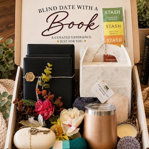 May include: A gift box labeled Blind Date with a Book containing black wrapped items with gold wax seals, a white box with floral patterns, Stash tea bags, a rose gold tumbler, a white soap bar with a key charm, and a small candle in a tin.