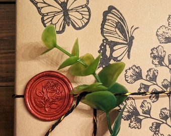 May include: A tan rectangular box decorated with black ink stamps of two butterflies and a fern-like plant. A red wax seal featuring a rose design is attached to the box, which is tied with black and gold twine and adorned with a green sprig.