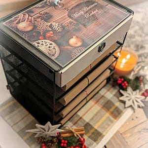 May include: A black metal mesh desktop bookshelf with four tiers. The top surface features a printed wood-grain design with Christmas ornaments, pinecones, and the text Merry Christmas Rachael. A green wax seal is visible on the front edge.