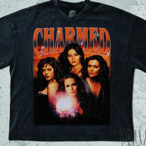 May include: Dark gray t-shirt featuring the word "CHARMED" in orange lettering above a graphic of four women. The graphic has a vintage, distressed look, with a faded orange and red background.