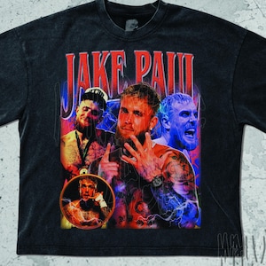 May include: Black t-shirt featuring a graphic of Jake Paul with red text that reads "JAKE PAUL". The design includes multiple images of Jake Paul with blue and red accents. The shirt has a vintage, distressed look.