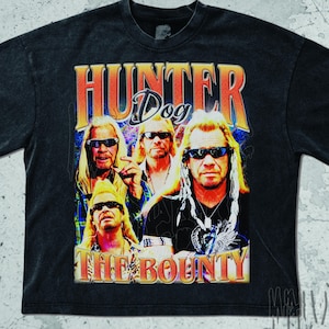 May include: Dark gray t-shirt featuring a graphic of four people with sunglasses and long blonde hair. The text "HUNTER Dog THE BOUNTY" is printed in orange and yellow. The shirt has a vintage, distressed look.