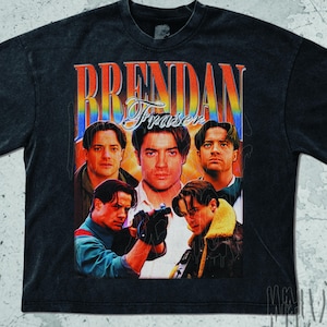 May include: Dark grey t-shirt with a retro graphic of Brendan Fraser. The design features multiple images of the actor with the name "Brendan Fraser" in a vintage font. The shirt has a faded, worn appearance.