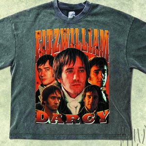 May include: A gray t-shirt featuring a graphic of several portraits of actors with the text "FITZWILLIAM DARCY" in orange and white. The shirt has a vintage, distressed look.