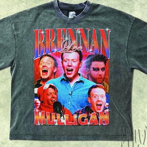 May include: A gray t-shirt featuring a vintage-style graphic of Brennan Lee Mulligan and other people. The graphic has the text "Brennan Lee Mulligan" in a retro font. The shirt has a crew neck and short sleeves.