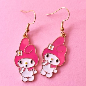 May include: A pair of gold dangle earrings featuring a pink and white cartoon character with a flower on her head. The character is wearing a pink hat and a white dress.