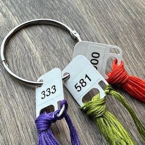 May include: Three embroidery floss spools with color numbers 333, 581, and 300 attached to a silver ring. The floss colors are purple, green, and orange.