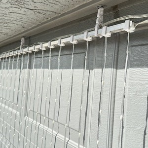 May include: White string lights hanging from a white metal track attached to the side of a garage. The lights are arranged in a grid pattern and are hanging down in long strands.