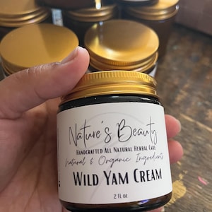 May include: A jar of Nature's Beauty Wild Yam Cream, 2 fl oz, held in a hand. The label reads "Handcrafted All Natural Herbal Care, Natural & Organic Ingredients." The jar has a gold lid and a brown glass body. Other jars are visible in the background.