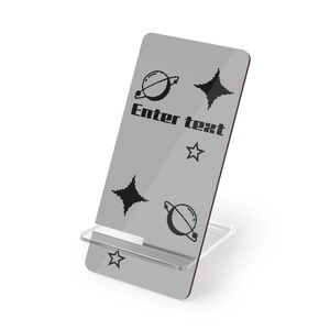 May include: A gray phone stand with a clear plastic base. The stand has a space holder for a phone and features a design of planets and stars. The text "Enter text" is printed on the stand.