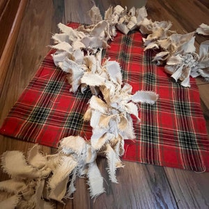 May include: A decorative garland made of off-white, frayed fabric strips arranged in a winding pattern over a rectangular red, black, and green plaid fabric cloth. The fabric strips have raw, uneven edges and are tied together in a continuous chain.