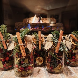 May include: Five glass jars filled with cranberries, dried orange slices, and greenery, topped with cinnamon sticks and burlap. The jars are arranged on a brick surface in front of a fireplace with a burning fire.