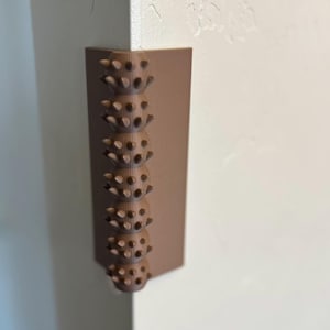 Puede incluir: Un protector de esquina marrón con una superficie texturizada. El protector tiene una serie de protuberancias elevadas que crean una superficie táctil.