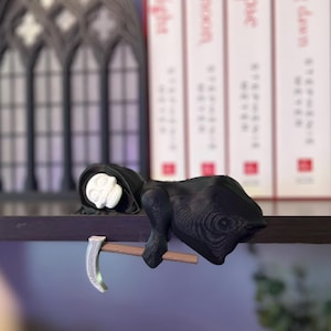 May include: A black Grim Reaper figurine with a white face and a scythe. The figurine is resting on a dark shelf, with books and a decorative gothic window in the background. The figurine is a unique decorative item.