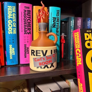May include: A tan jug with a handle and a flame-shaped topper. The jug features the text "REV UP" in black and "Carl's Jug O' BOOM" in red on a grey rectangular graphic that resembles duct tape, with black "XXX" markings at the bottom.