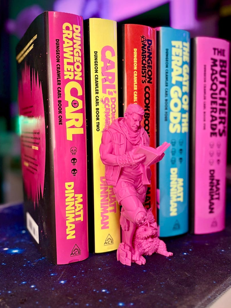 May include: A collection of colorful books from the "Dungeon Crawler Carl" series, with a pink figurine of a man reading. The book spines are pink, yellow, red, blue, and pink, with titles and author names in white and yellow.