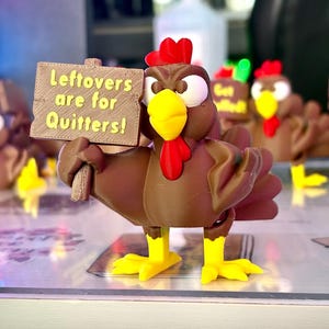 May include: A brown turkey figurine with a red wattle and comb, yellow beak and feet, holding a sign that reads "Leftovers are for Quitters!". Other turkey figurines are in the background.