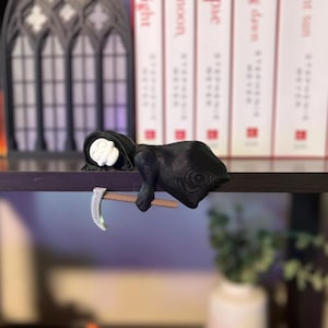 May include: A black Grim Reaper figurine with a white skull face and a scythe. The figurine is resting on a wooden shelf, with books and decorative elements in the background. The scythe is silver and brown.