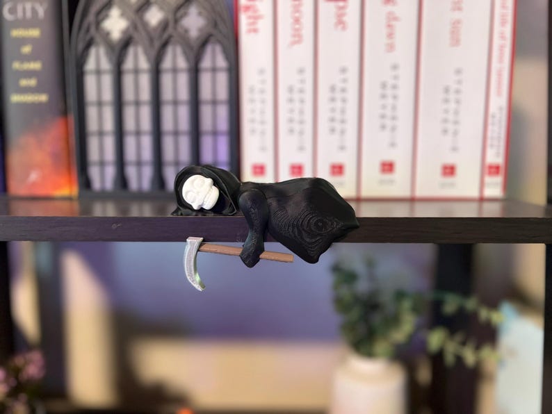 May include: A black Grim Reaper figurine with a white skull face and a scythe. The figurine is resting on a wooden shelf, with books and decorative elements in the background. The scythe is silver and brown.