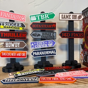 May include: A collection of novelty signposts with various phrases and words, including "Fantasy," "Thriller," and "Game On!" The signs are in various colors, mounted on black stands, and arranged on a wooden shelf. Some signs are lying flat.
