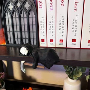 May include: A black Grim Reaper figurine with a scythe, resting on a shelf. The figurine has a white face and is wearing a black hooded robe. Books and a decorative window are in the background.