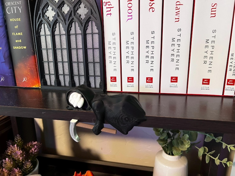 May include: A black Grim Reaper figurine with a scythe, resting on a shelf. The figurine has a white face and is wearing a black hooded robe. Books and a decorative window are in the background.