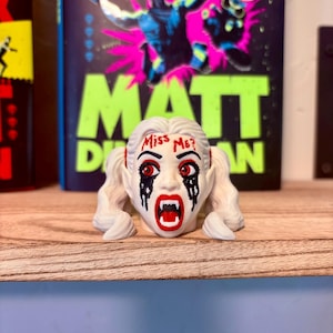 May include: A white figurine head with blonde pigtails, red eyes, and black paint dripping from the eyes. The forehead features the red text "Miss Me?" and the mouth is open, revealing white fangs against red lips, creating a vampire-like appearance.
