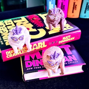 K&ouml;nnte beinhalten: Drei skurrile Katzenfiguren mit Einhornh&ouml;rnern und Brillen liegen auf zwei B&uuml;chern. Die B&uuml;cher tragen den Titel "Dungeon Crawler" und den Namen des Autors "Matt Dinniman" in leuchtenden Farben. Der Hintergrund umfasst bunte Textmarker.