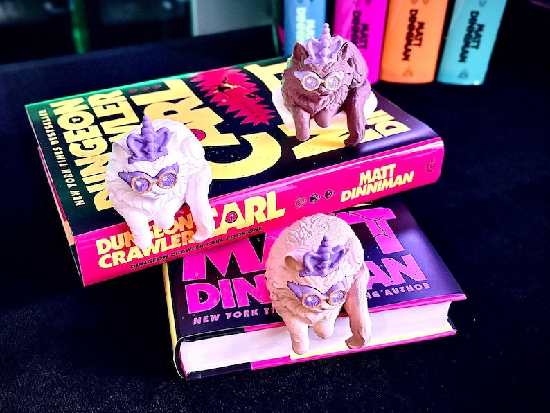 K&ouml;nnte beinhalten: Drei skurrile Katzenfiguren mit Einhornh&ouml;rnern und Brillen liegen auf zwei B&uuml;chern. Die B&uuml;cher tragen den Titel "Dungeon Crawler" und den Namen des Autors "Matt Dinniman" in leuchtenden Farben. Der Hintergrund umfasst bunte Textmarker.