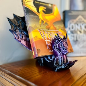 May include: A purple and black dragon bookend holding a book titled "Wings of Fire: The Eight  Dragons".